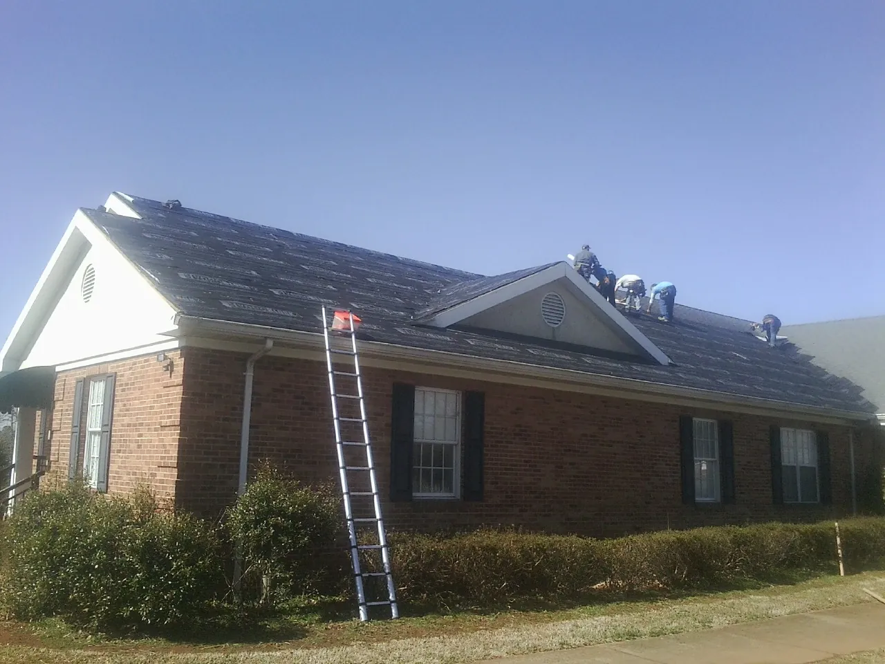 Crew tearing shingles off a brick ranch roof with a ladder set — Emergency Roof Repair work by our crew in Cataula