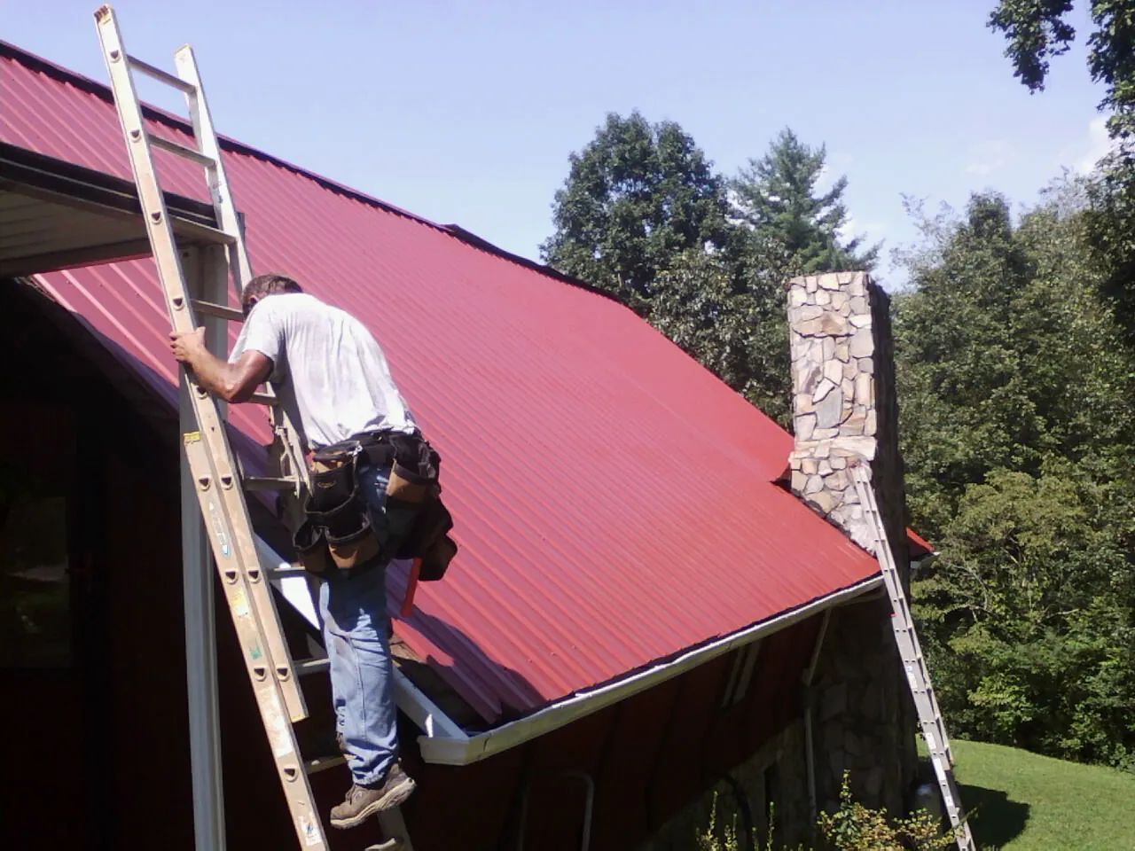Roofer on a ladder beside a red metal roof and stone chimney — Soffit & Fascia Repair by Roofing Brothers in Cataula